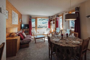 A cozy room with a sofa, a TV, a dining table set for four, and a window with a view of a snowy landscape. - Alpine Apartment with Parking - on slopes (Méribel)