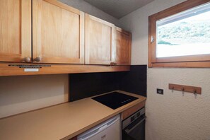 A compact kitchen with wooden cabinets, a black stove, and a window with a view of a snowy landscape. - Alpine Apartment with Parking - on slopes (Méribel)