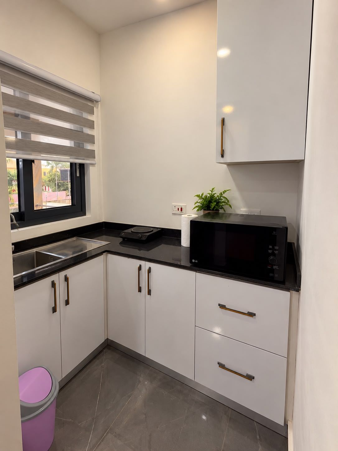 A modern kitchen with white cabinets, a black countertop, a microwave, and a window with blinds.