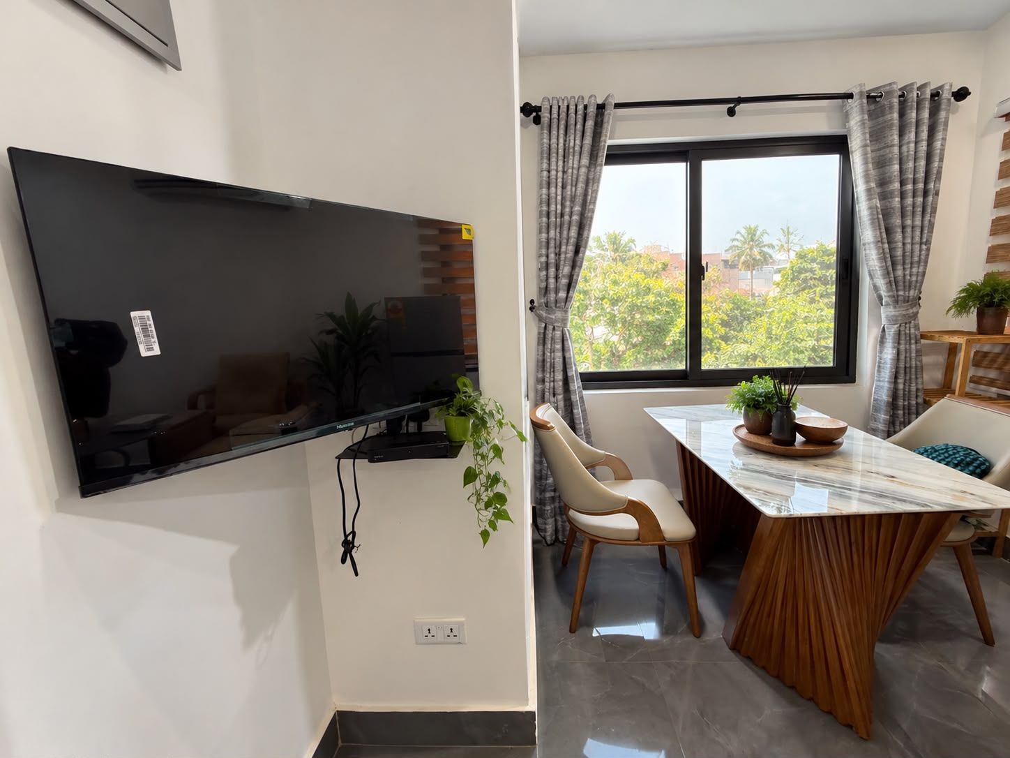 Un espace de vie moderne doté d’un téléviseur à écran plat fixé au mur, d’un coin repas avec une table et des chaises, et d’une fenêtre avec des rideaux.