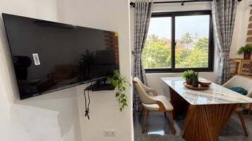 Un espace de vie moderne doté d’un téléviseur à écran plat fixé au mur, d’un coin repas avec une table et des chaises, et d’une fenêtre avec des rideaux.