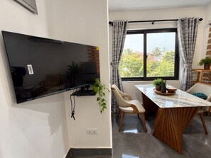 Un espace de vie moderne doté d’un téléviseur à écran plat fixé au mur, d’un coin repas avec une table et des chaises, et d’une fenêtre avec des rideaux.