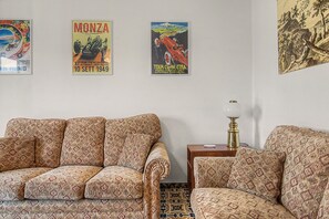 Un saló amb sofàs estampats, una taula petita amb una làmpada i tres cartells vintage de cotxes a la paret.