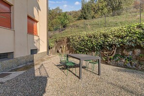 A gravel patio with a table and chairs, a building with a window and shutters, and a hedge with climbing plants. - Apartment 'Aurora Alta' with Balcony, Wi-Fi and Air Conditioning (Maslianico)