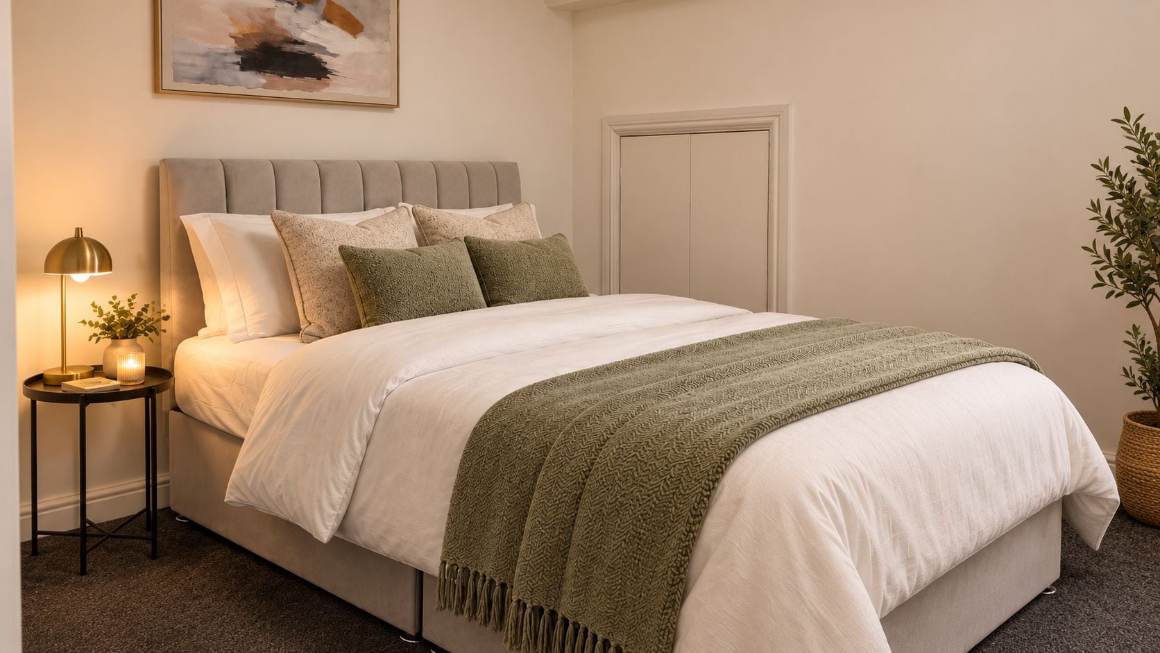 A neatly made bed with a grey headboard, a bedside table with a lamp, and a potted plant.
