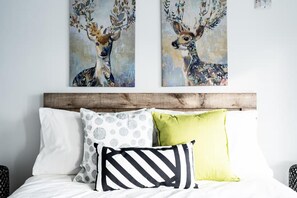 Een bed met witte lakens, een kussen met dessin, een gestreept kussen en een effen groen kussen. Daarboven hangen twee schilderijen van herten op de muur.