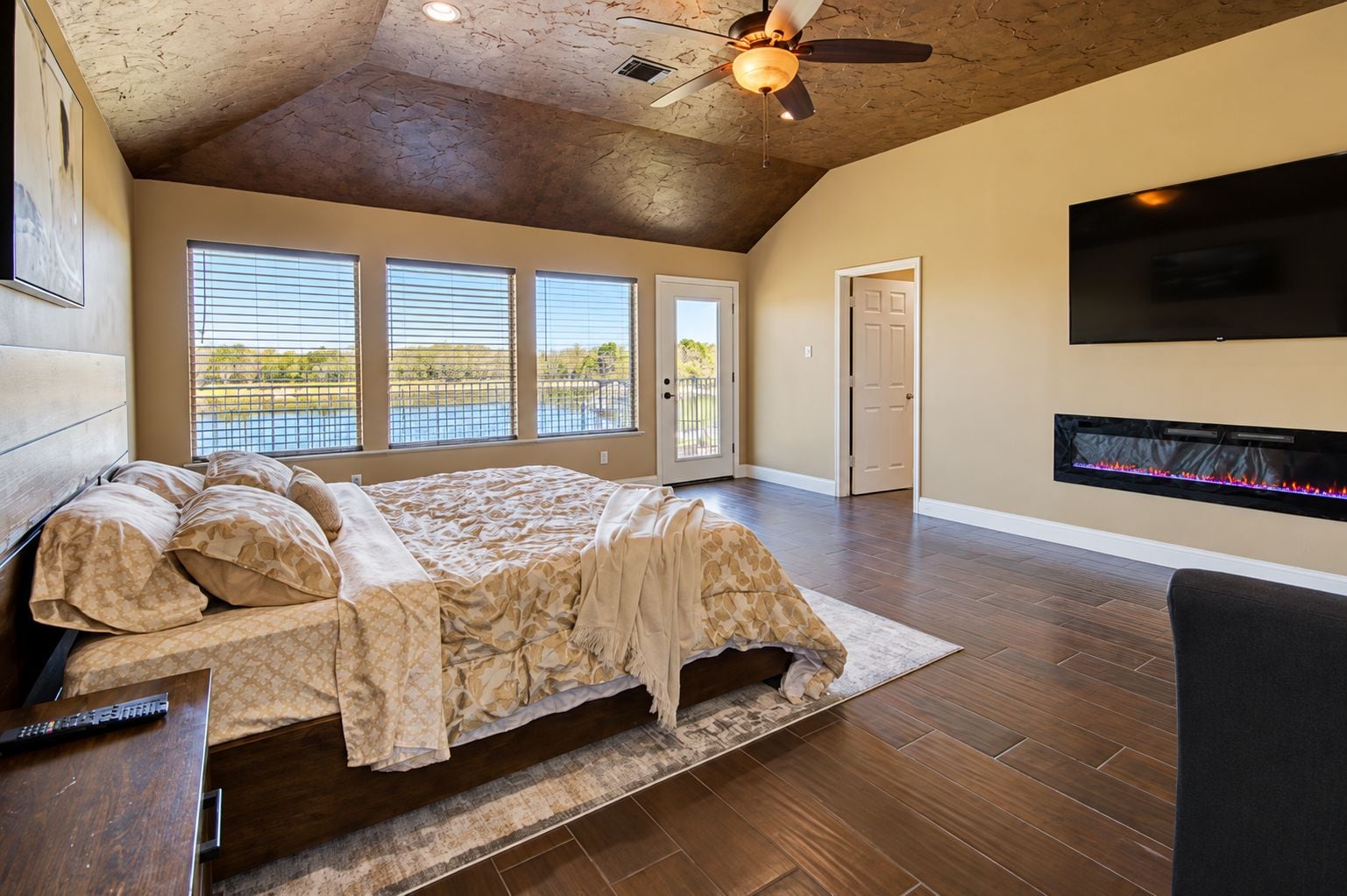 A spacious bedroom with a large bed, a fireplace, a ceiling fan, and a view through the windows.