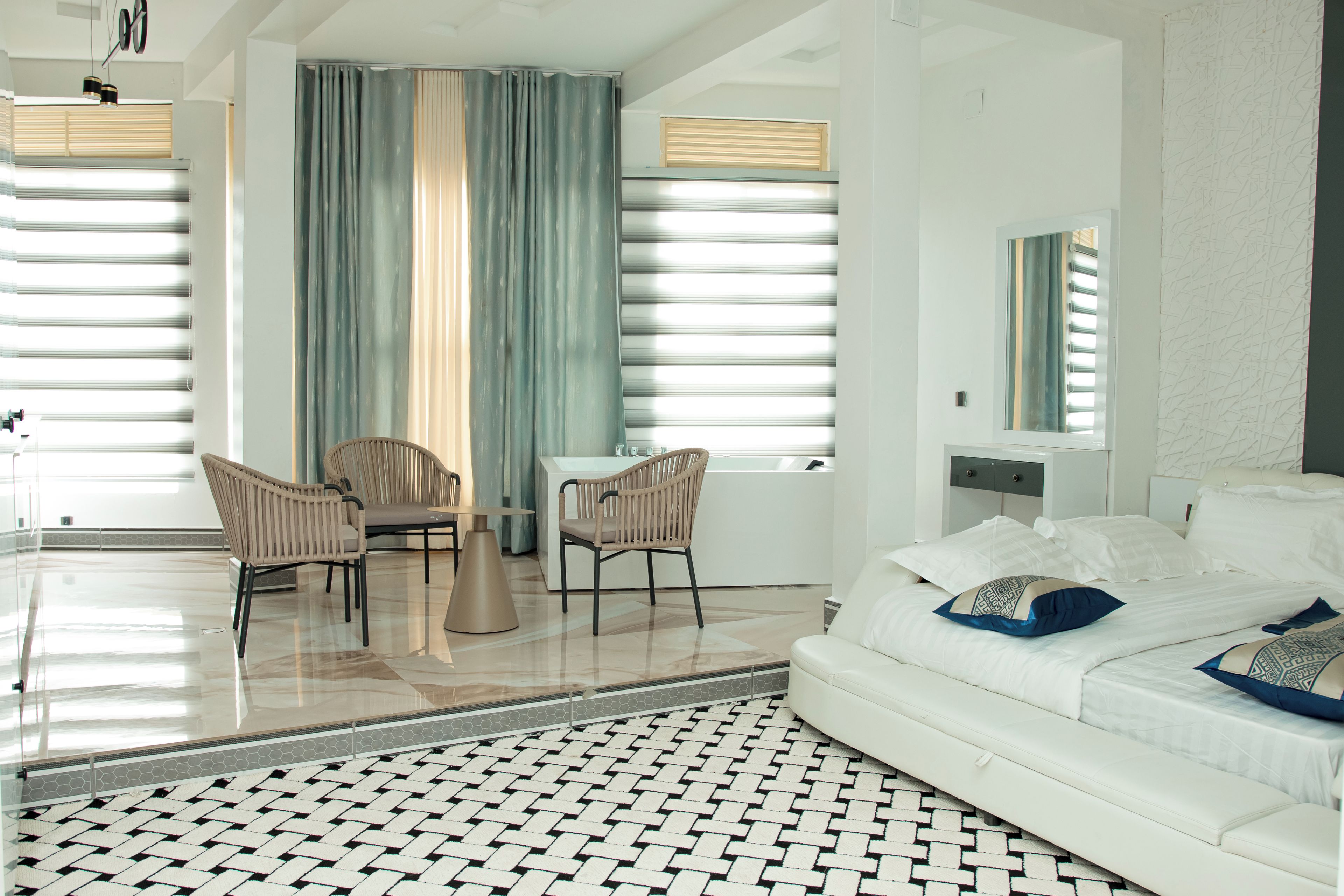 Ein modernes Hotelzimmer mit einem großen Bett, einer Sitzecke mit zwei Stühlen, einem kleinen Tisch und einem großen Fenster mit Vorhängen.