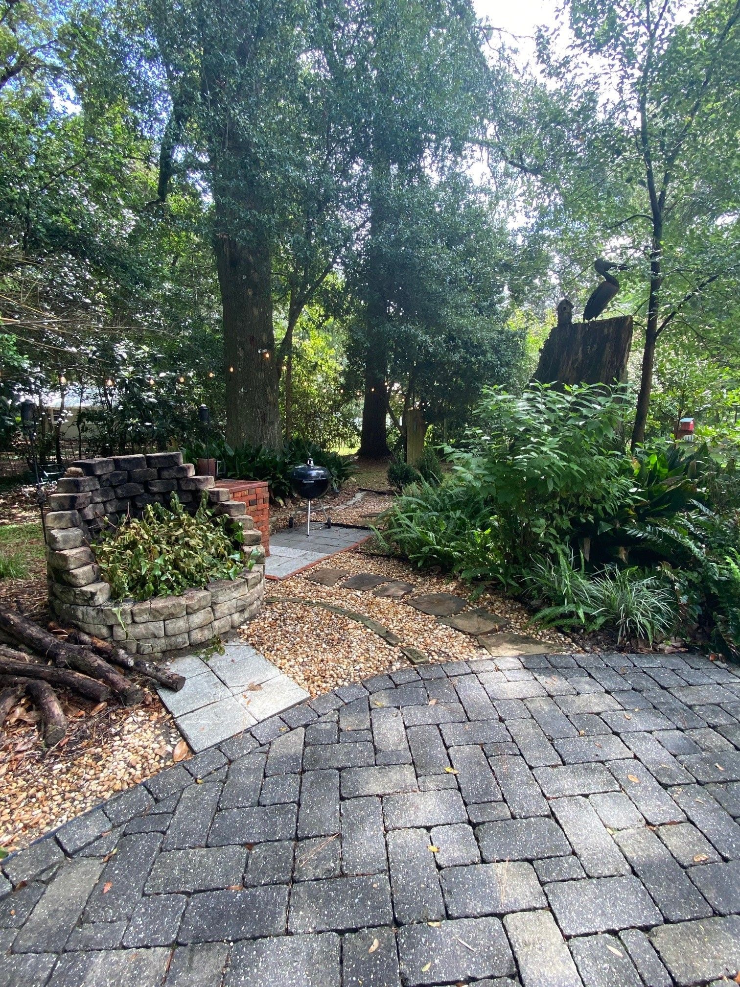 A paved patio area with a brick wall and a grill, surrounded by lush greenery and trees.