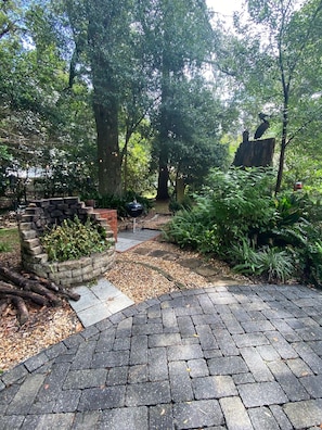 A paved patio area with a brick wall and a grill, surrounded by lush greenery and trees.