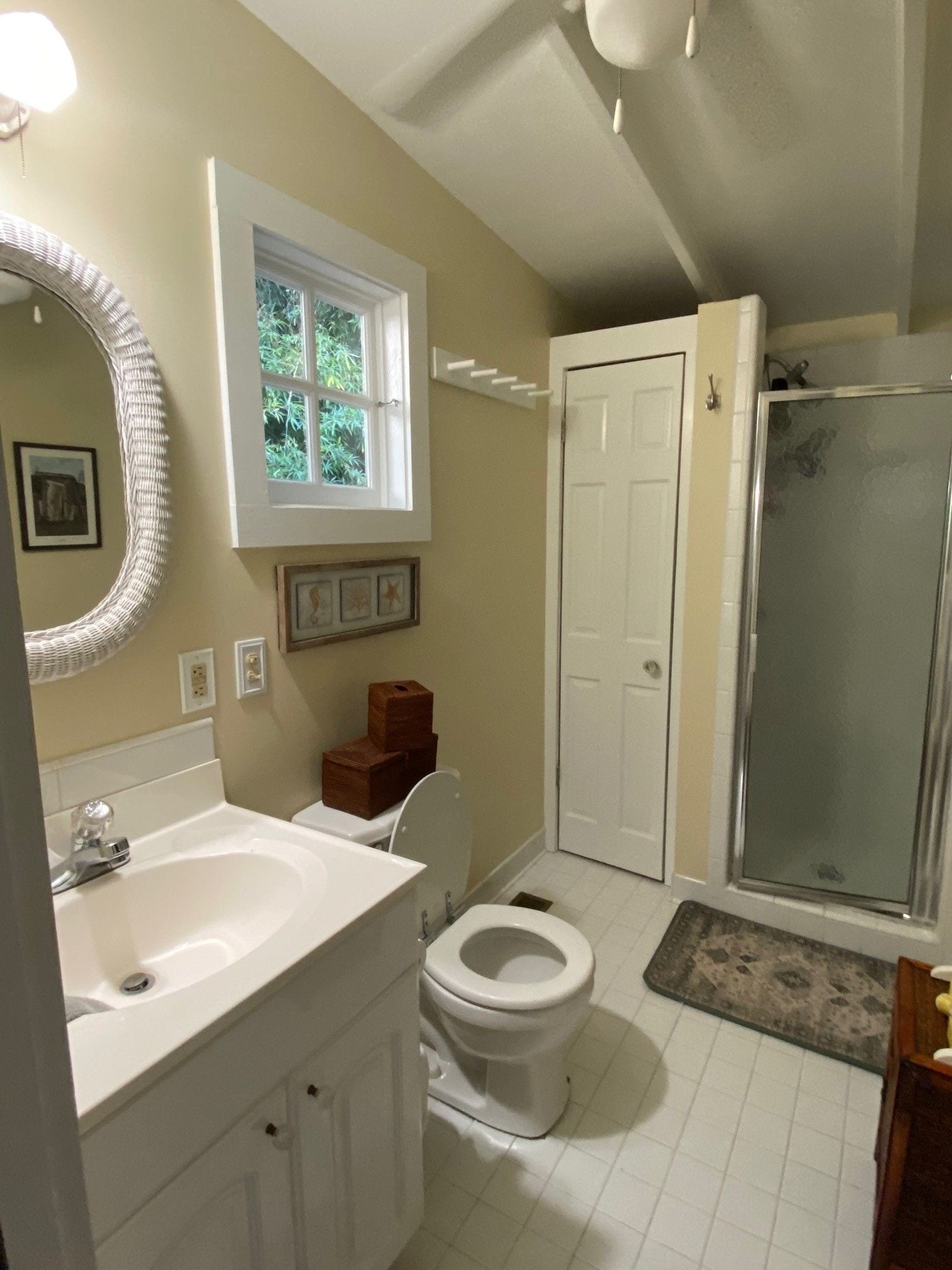 A bathroom with a white sink, toilet, and shower. There is a window above the sink and a door leading to another room.