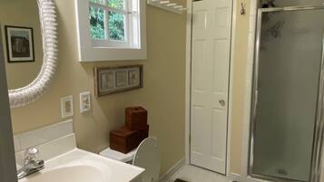 A bathroom with a white sink, toilet, and shower. There is a window above the sink and a door leading to another room.