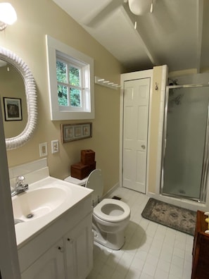 A bathroom with a white sink, toilet, and shower. There is a window above the sink and a door leading to another room.
