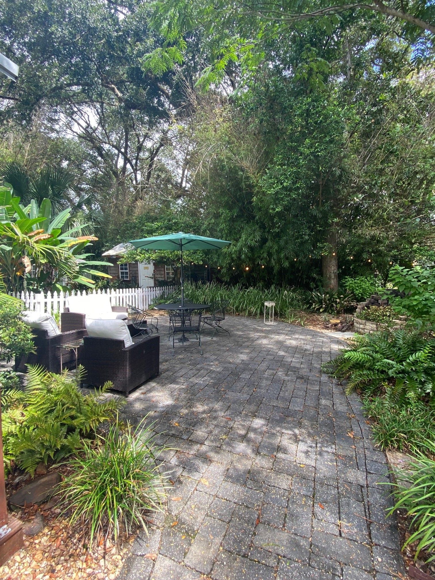 A paved patio area with outdoor furniture and a table under a green umbrella, surrounded by lush greenery.