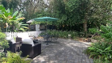 A paved patio area with outdoor furniture and a table under a green umbrella, surrounded by lush greenery.