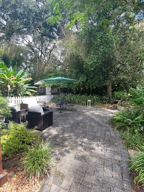 A paved patio area with outdoor furniture and a table under a green umbrella, surrounded by lush greenery. - Cozy 1-bedroom cottage in charming Magnolia Springs with all the amenities (Magnolia Springs)