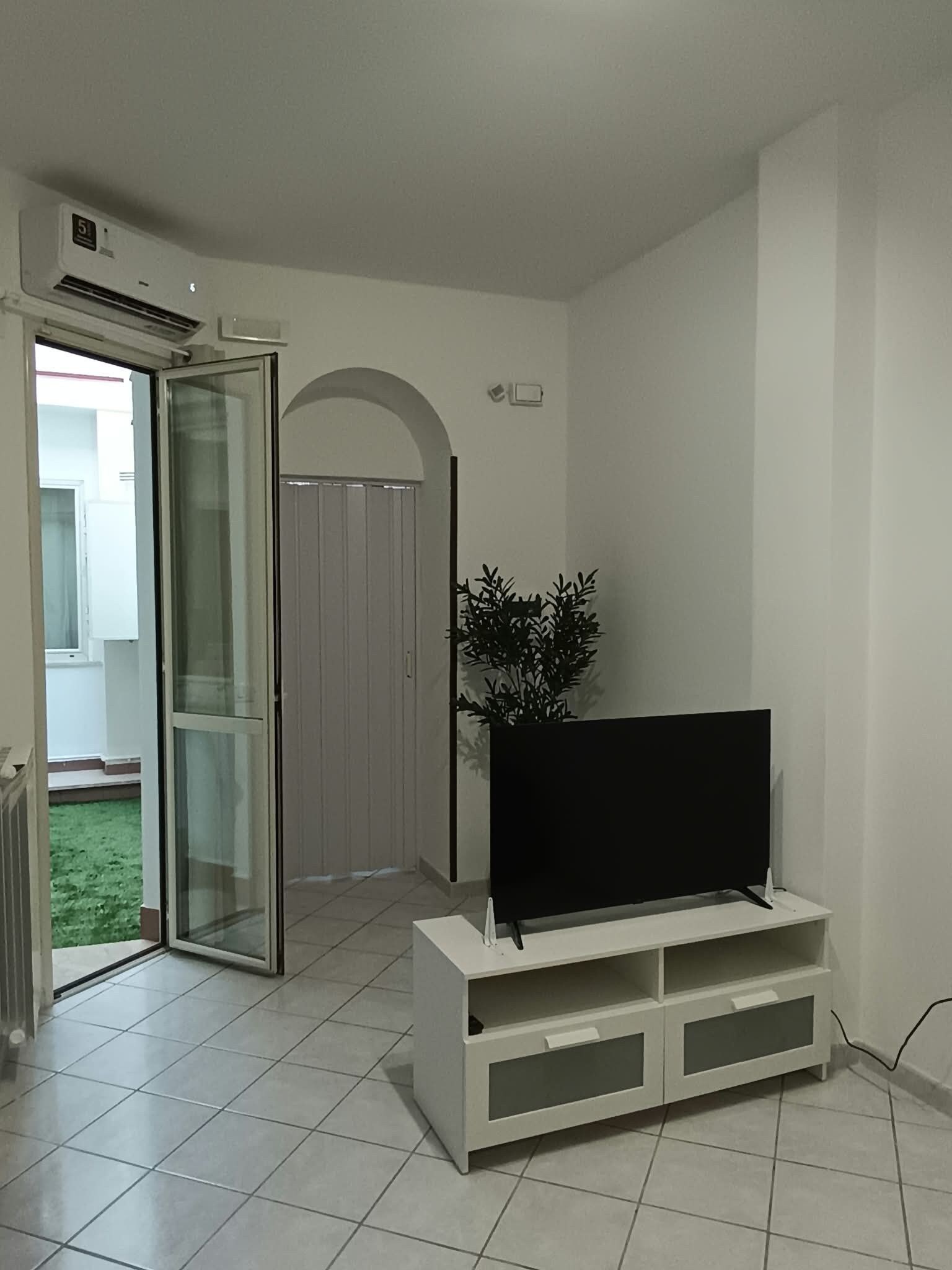 Un soggiorno con una TV a schermo piatto su un mobile bianco, una porta vetrata scorrevole che dà su un balcone e una pianta in vaso.