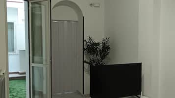 Un soggiorno con una TV a schermo piatto su un mobile bianco, una porta vetrata scorrevole che dà su un balcone e una pianta in vaso.