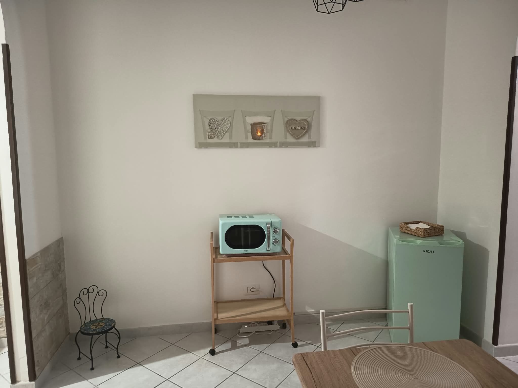 Una piccola angolo cottura con un forno a microonde color acquamarina su un supporto in legno, un tavolino con tovaglietta intrecciata e un armadietto verde con un cesto sopra.