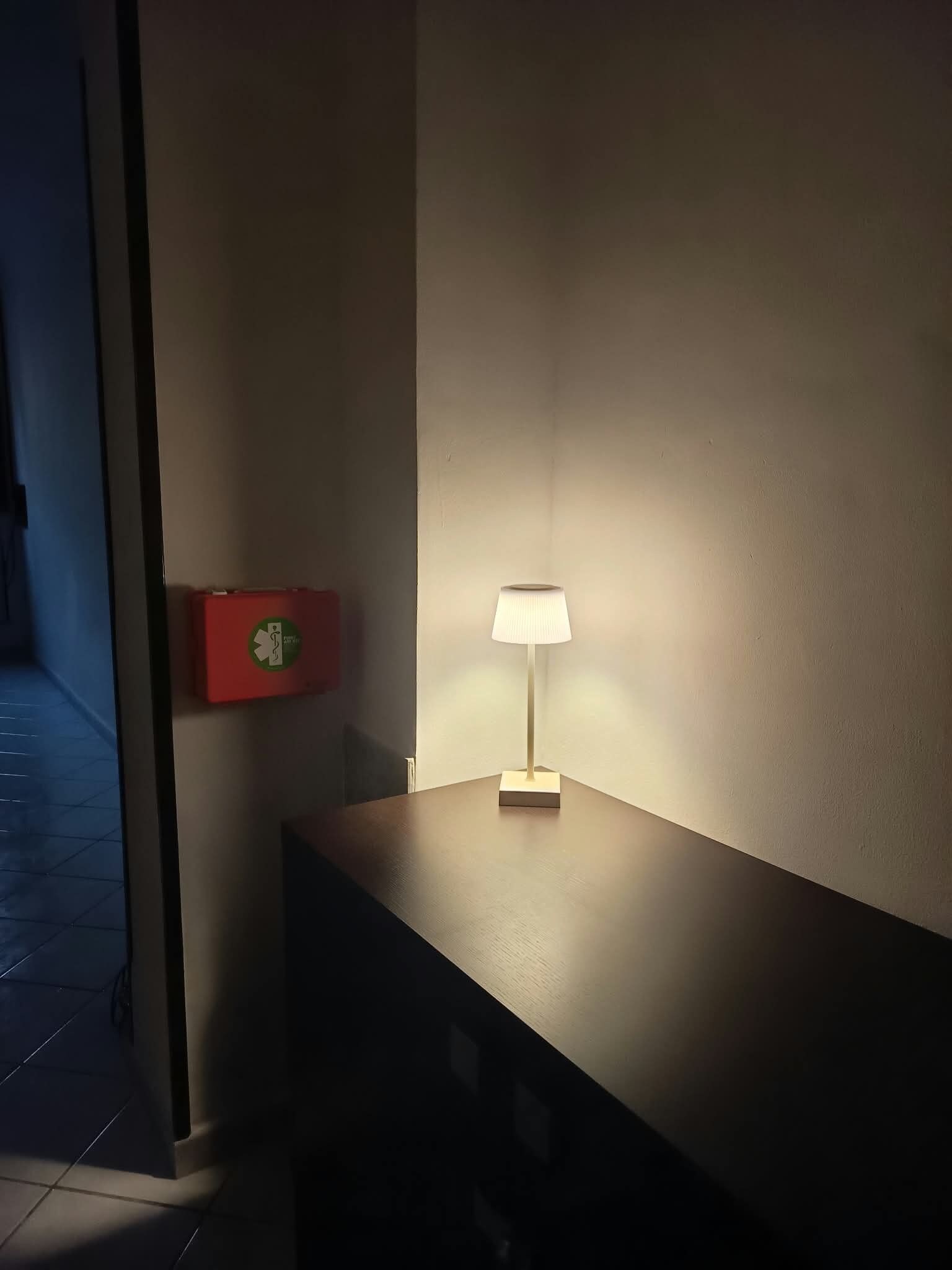 Sängyn vieressä oleva yöpöytä, jossa on lamppu, punainen hätäuloskäynti-opaste ja seinälle kiinnitetty kello.