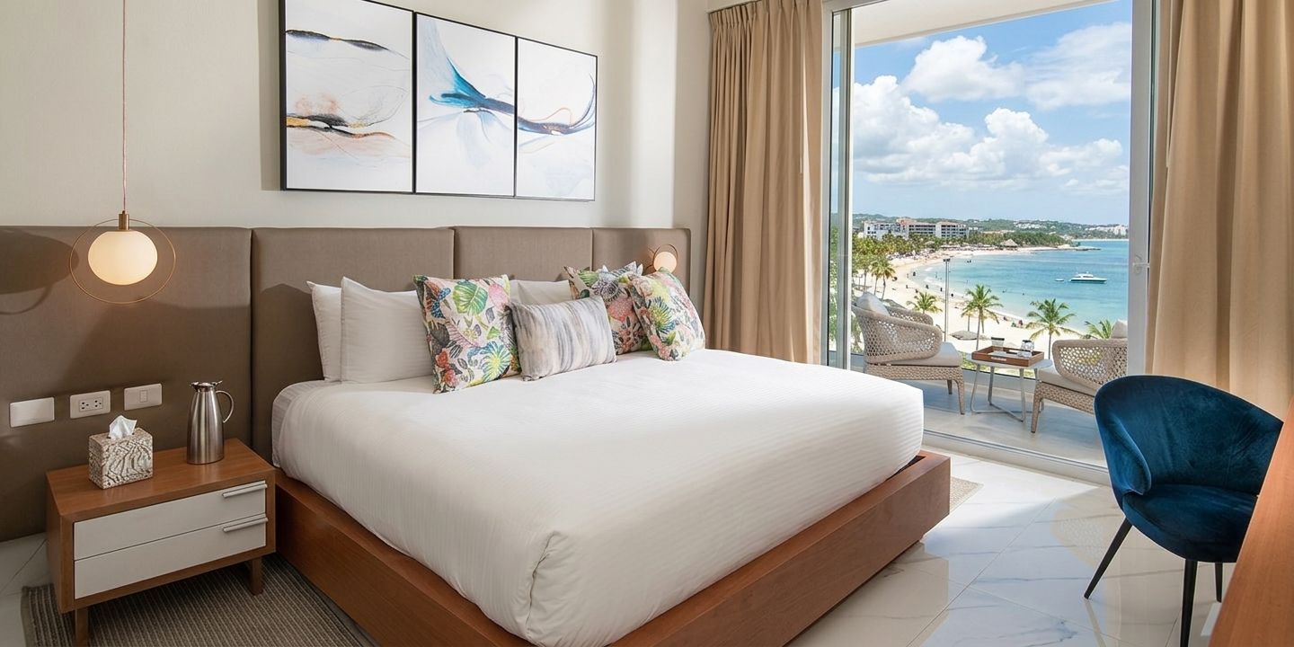 A hotel room with a large bed, a nightstand with a lamp, a view of the beach and ocean through the window.