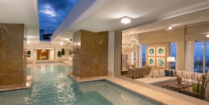 A modern indoor swimming pool with a view of the city at dusk, a dining area with a table and chairs, and a living area with a sofa and a lamp. - Sunrise Suites All Inclusive (Puerto Plata)