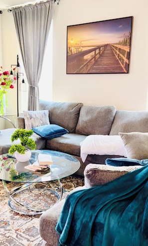 Un salon comprenant un canapé, une table basse en verre, un tapis et un tableau représentant un coucher de soleil sur un pont.