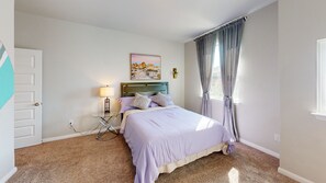 A bedroom with a bed, a bedside table with a lamp, a painting on the wall, and a window with curtains. -  Cozy Comfort Home near Randolph AFB, Fort Sam& I 10 -Spacious 5-bedroom house (Converse)