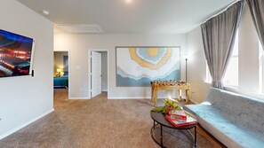 A living room with a sofa, a foosball table, a painting, and a TV displaying a cityscape. -  Cozy Comfort Home near Randolph AFB, Fort Sam& I 10 -Spacious 5-bedroom house (Converse)
