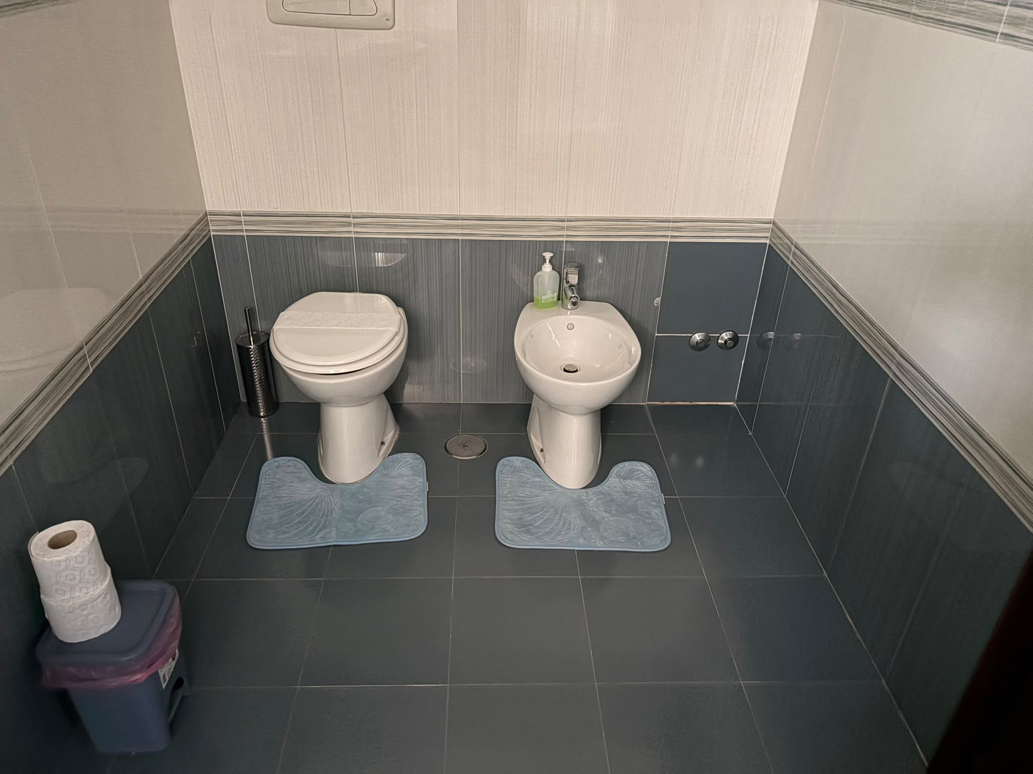 Un baño pequeño con dos inodoros, un lavamanos y un cubo de basura.