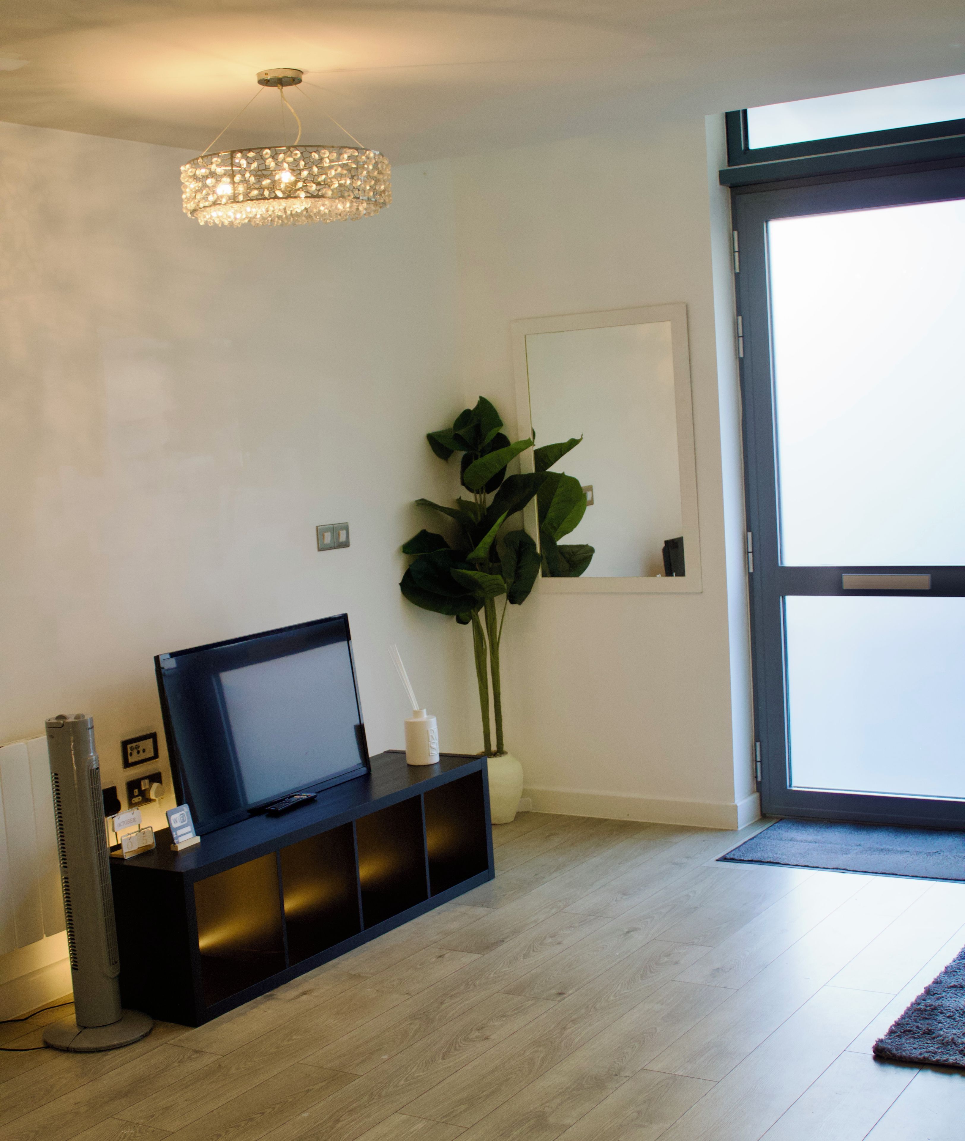 Una sala de estar moderna con un televisor de pantalla plana sobre un soporte negro, una planta grande en maceta, una lámpara araña decorativa y un puerta corrediza de vidrio que da a un balcón.