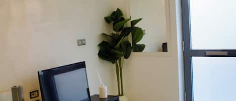 Una sala de estar moderna con un televisor de pantalla plana sobre un soporte negro, una planta grande en maceta, una lámpara araña decorativa y un puerta corrediza de vidrio que da a un balcón.