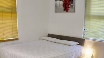 Een slaapkamer met een bed, nachtkastje, lamp, schilderij aan de muur en een raam met jaloezieën.