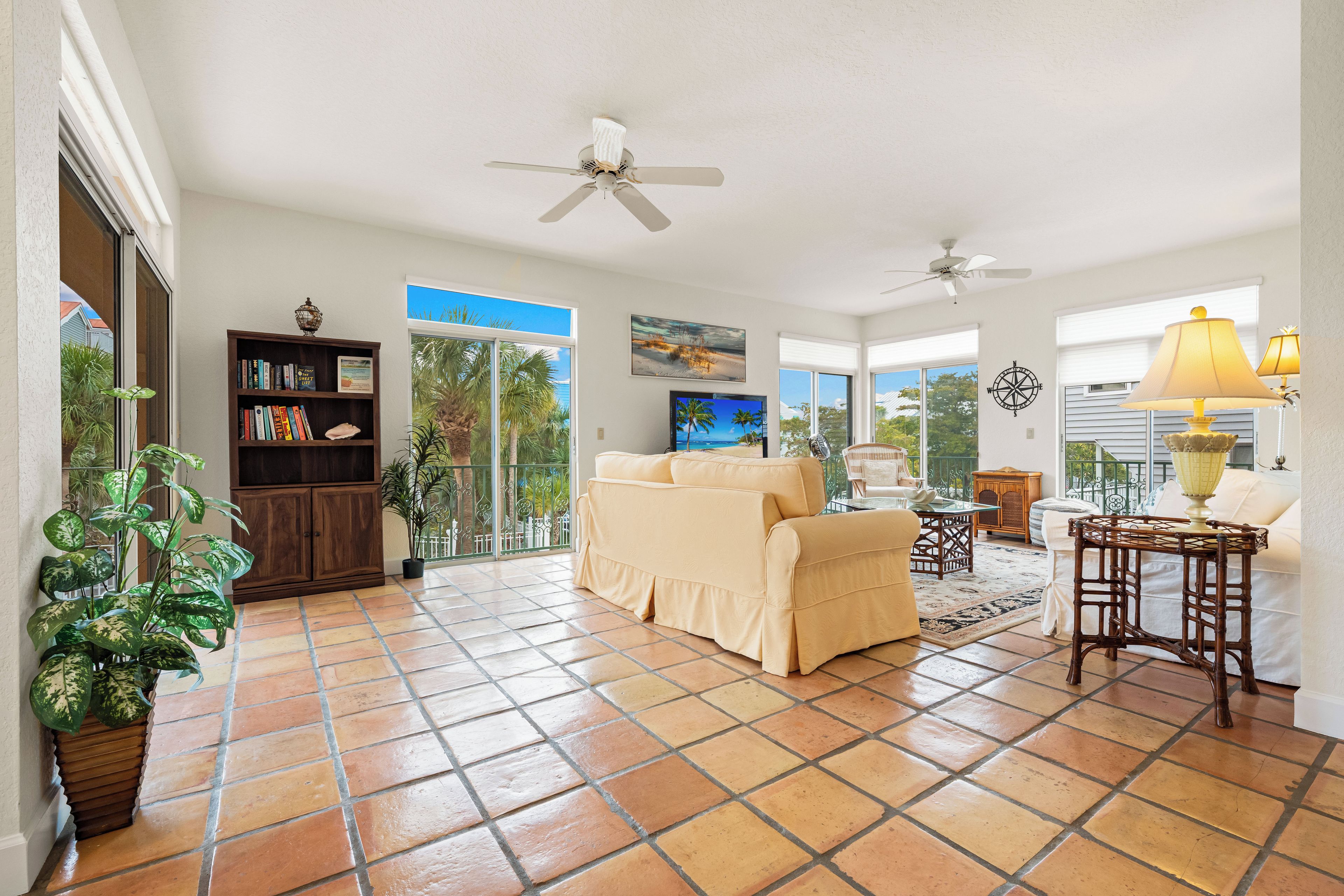 A spacious living room with a beige sofa, a wooden bookshelf, and a large window with a view of palm trees.