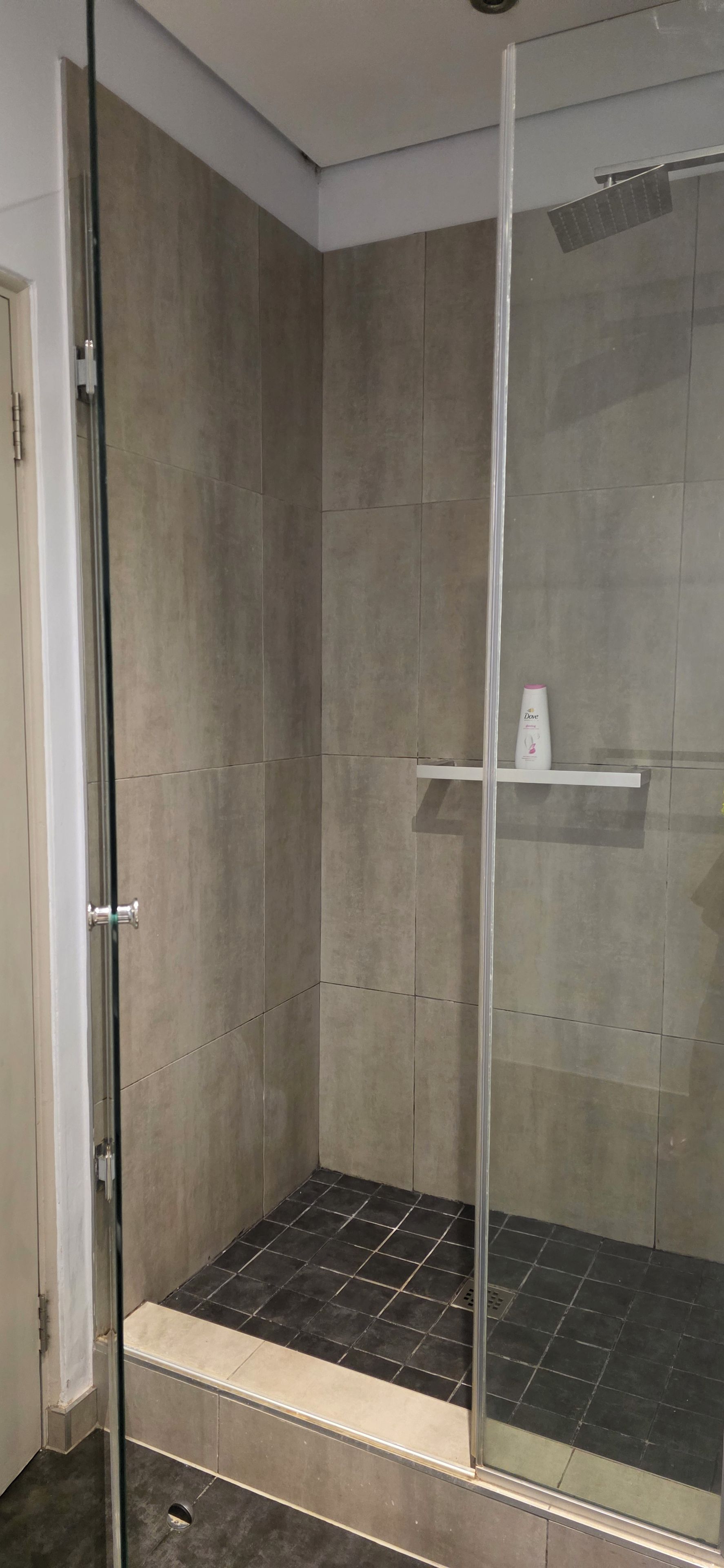 Zona de ducha con paredes de vidrio, piso de baldosas y un estante con artículos de tocador.