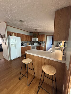 A kitchen with wooden cabinets, a white refrigerator, a microwave, a stove, and two stools. - Waterfront Sunset Home on Lyman Lake Private Dock Hot Tub Sleeps 8 Near GSP (Lyman)