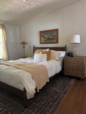 Un lit bien fait, avec une couette blanche et un plaid beige, une table de chevet en bois avec une lampe, un tableau encadré au mur et un tapis à motifs sur le sol.
