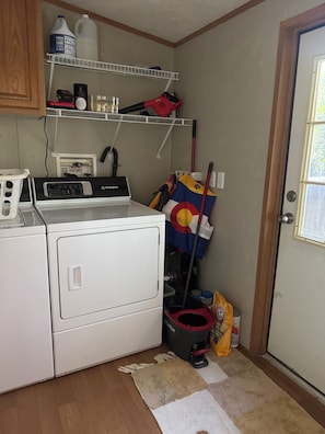 A laundry area with a front-loading washer, a shelf with cleaning supplies, a mop, and a door with a window. - Waterfront Sunset Home on Lyman Lake Private Dock Hot Tub Sleeps 8 Near GSP (Lyman)