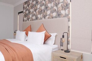 Una cama bien hecha con almohadas blancas y naranjas, un plaid marrón claro y una moderna mesita de noche con una lámpara.