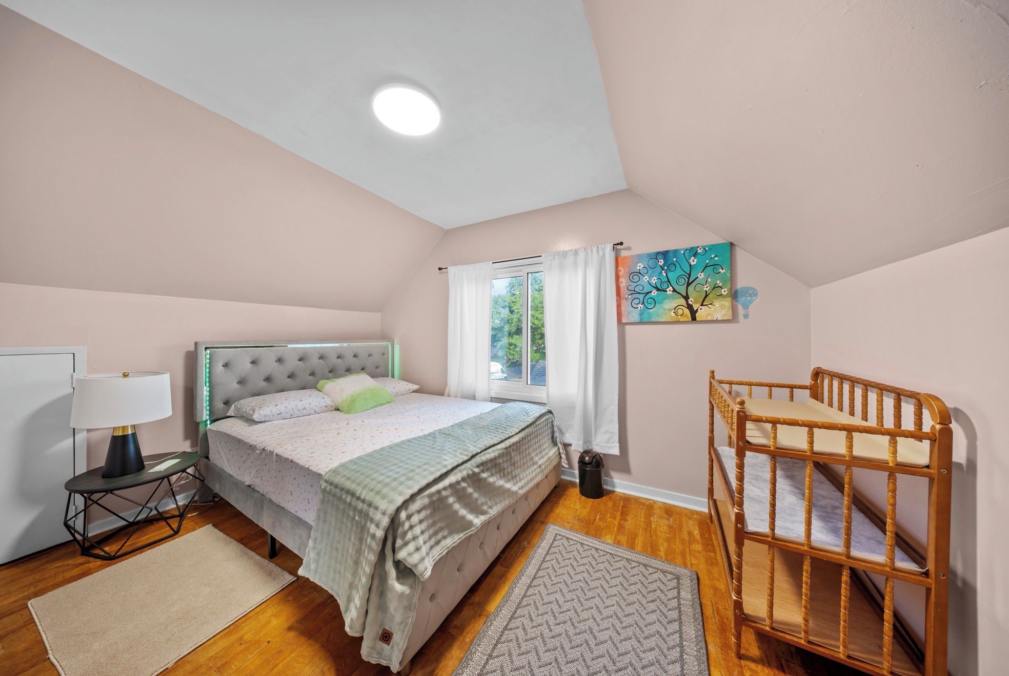 A bedroom with a bed, a nightstand, a lamp, a crib, and a window with curtains.