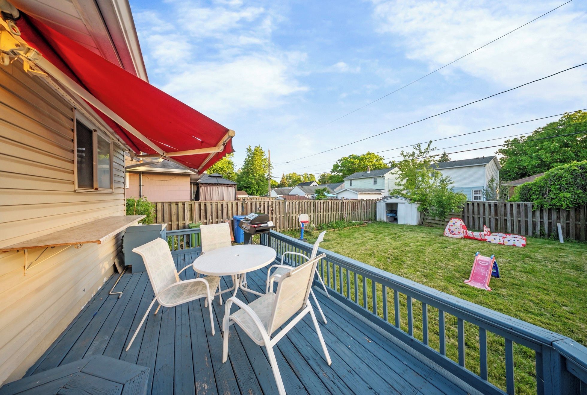 A deck with outdoor furniture and a red awning, overlooking a backyard with a wooden fence and a slide.