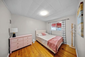 Een slaapkamer met een bed, een dressoir, een lamp, een raam met gordijnen en een kapstok.