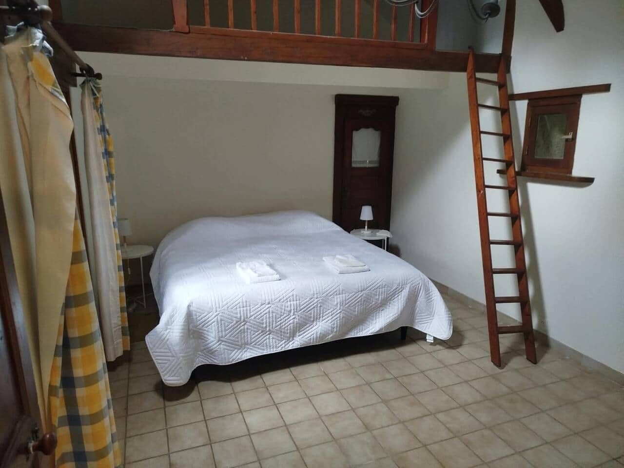 Habitación con cama individual, una escalera de madera, una mesita de noche pequeña y una ventana con marco de madera.