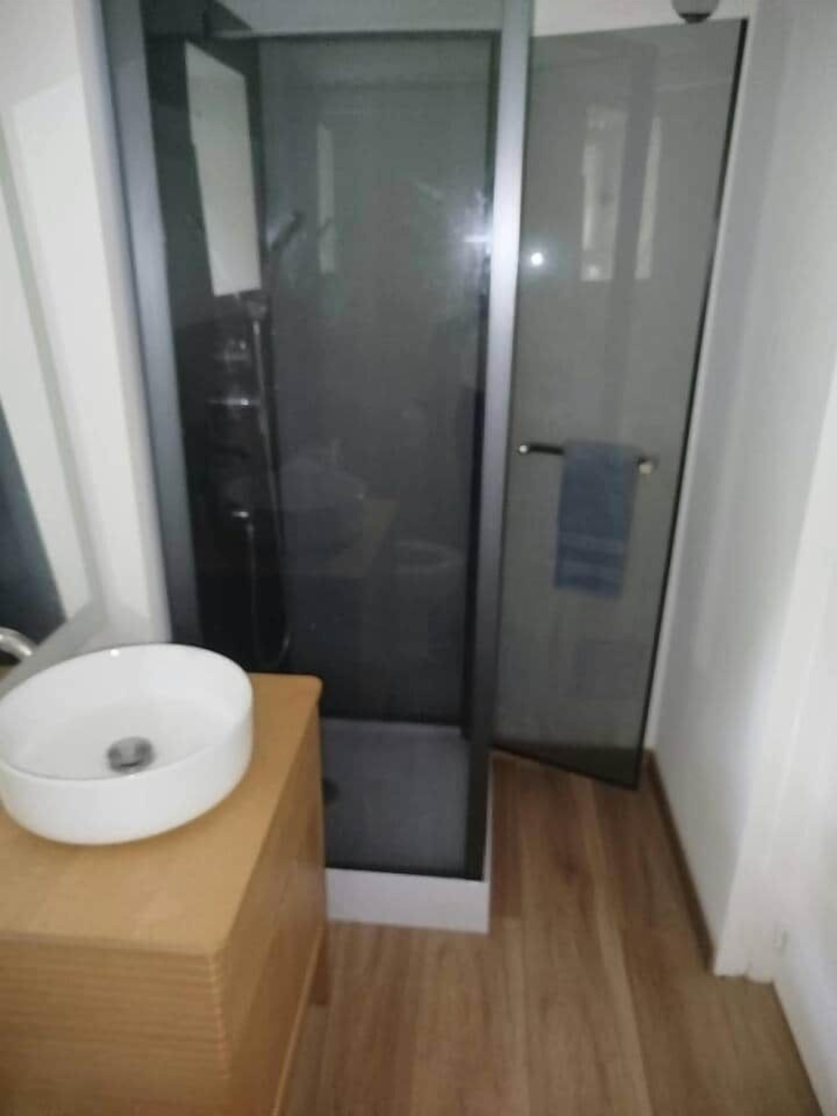 Un baño con cabina de ducha de vidrio, un lavamanos blanco sobre un mueble de madera y una toalla colgada en una puerta.