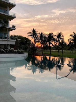 A modern building with a reflective pool, palm trees, and a sunset in the background. - Luxury 2BR Condo - Marina & Golf Views, Resort Pool, Gym & Jacuzzi (Puerto Vallarta)