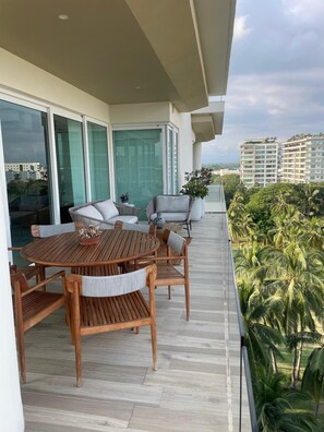 Uma varanda com mesa e cadeiras de madeira, uma porta de vidro que dá para um quarto e vista para palmeiras e edifícios.