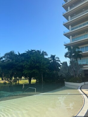 Uma piscina de borda curva, rodeada por palmeiras e um edifício com várias andares e varandas.