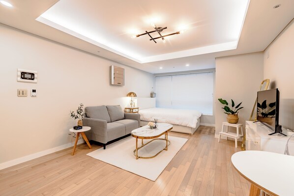 Una sala de estar moderna con un sofá, una mesa de centro, una mesita auxiliar con un reloj, una mesita de noche con una lámpara y un ventanal con persianas.