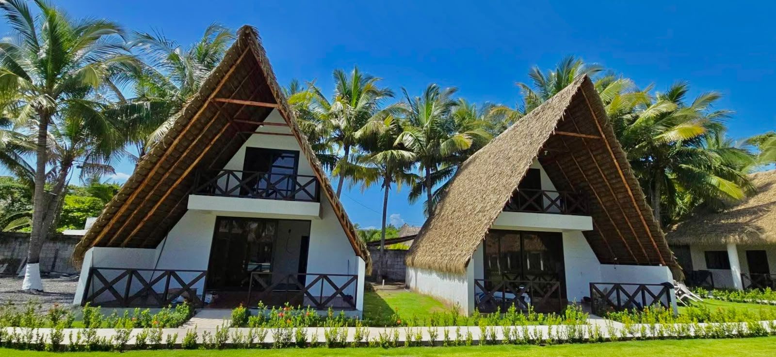 Deux bungalows aux toits de chaume, dotés de balcons, entourés de palmiers et d’une pelouse bien entretenue.