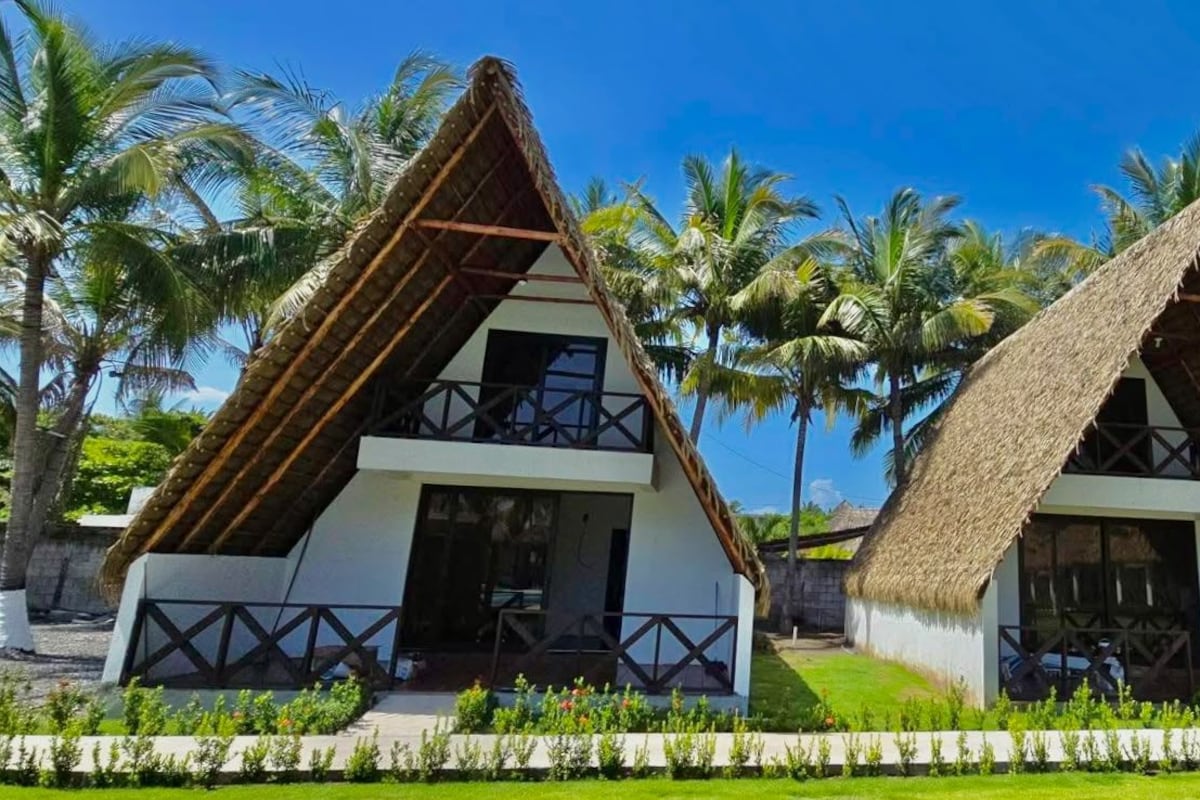 Two thatched-roof bungalows with balconies, surrounded by palm trees and a well-maintained lawn.